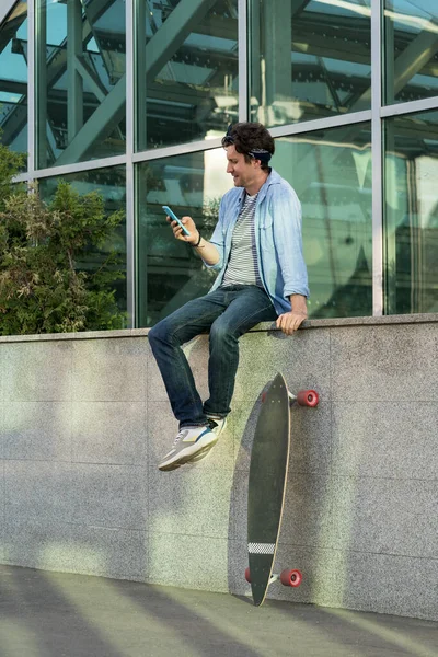Хіпстерський ковзаняр за допомогою смартфона тримає довгу дошку в руці, читаючи повідомлення в мобільному телефоні онлайн — стокове фото