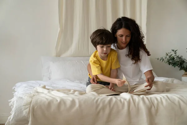 어린 어머니가 아침에 침대에 앉아 잠옷 차림으로 어린 아들 과 함께 책을 읽고 있다 — 스톡 사진