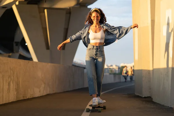 Νεαρή ανέμελη γυναίκα απολαμβάνει την οδήγηση skateboard στο ηλιοβασίλεμα. Ευτυχισμένο κορίτσι σε μοντέρνα ρούχα στυλ δρόμου — Φωτογραφία Αρχείου