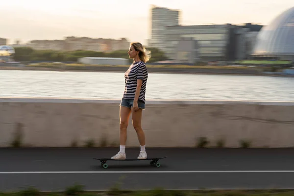 Femme décontractée d'âge moyen se détendre patiner sur longboard dans la soirée sur la ville floue le long de la rivière — Photo