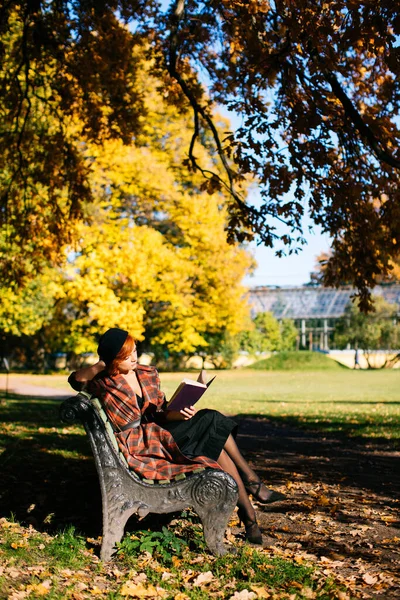Pelirroja con abrigo a cuadros y boina negra leyendo libro en el banco, descansando en el parque de otoño en el día soleado — Foto de Stock