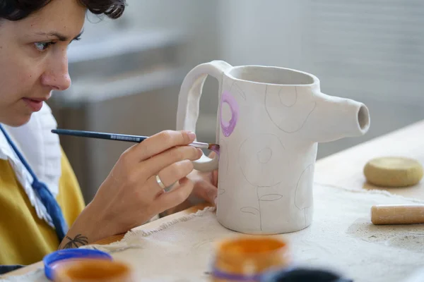 Художница держит кисть, создавая орнамент на глиняном кувшине в мастерской керамики. Керамическое рабочее место — стоковое фото