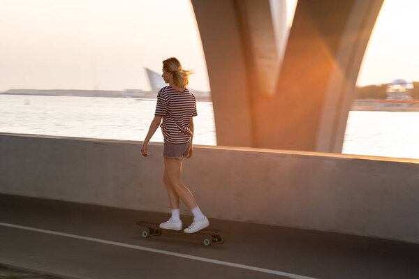 Woman skateboarder on longboard on street at sunset. Female longboarder riding alone alongside river