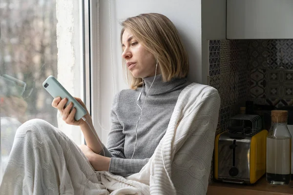 Uttråkad kvinna beroende och chatta i sociala medier, sitter på fönsterbräda hemma med mobiltelefon — Stockfoto