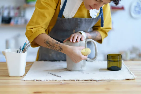 Estudio de cerámica en el interior: chica artista en delantal creando jarra de trabajo de arcilla húmeda sosteniendo herramientas profesionales — Foto de Stock