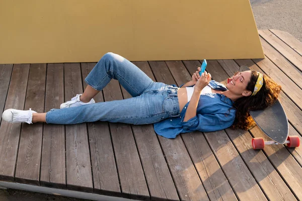 Güneş gözlüklü genç hippi kız dışarıda akıllı telefonuyla mesaj atıyor. Uzuntahtanın üzerinde rahatça yatıyor. — Stok fotoğraf