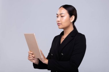 20 'li yaşlardaki Asyalı kadın portresinin yarısı siyah saçlı, resmi takım elbiseli. Ofisteki kadın gri arka planda dijital tablet gülümsemesi taşıyor.