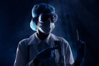 Stetoskop hijyen kaskı, lastik eldiven, şırınga, koruyucu yüz maskesi olan Asyalı doktor kadın. Bilim adamlarının elinde başarılı bir aşı olduğu görülüyor. koyu siyah duman arkaplanı