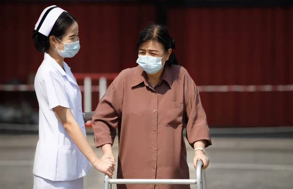 Nurse is helping Senior Elderly Asian Woman uses Aluminium Zimmer frame walker to support during rehabilitation to recovery. New Normal people with protective face mask under the Sun