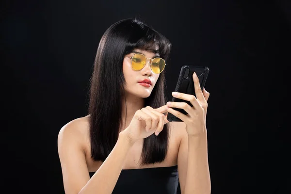 Fashion Beauty Woman has long straight black hair looks at camera and express feeling. Portrait of Asian girl use smart phone over black background, copy space
