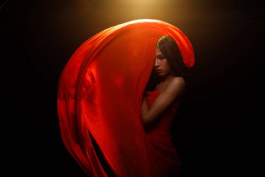 Moda güzeli kadının uzun siyah saçları var. Duygularını seksi bir şekilde ifade ediyor. Asyalı bir kızın portresi kırmızı elbiseler ve havada uçuşan kumaşlar siyah arka planda, boşluğu kopyala