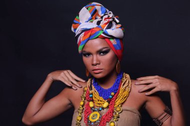 20 'li yaşlarda, Afrika Tepesi Kabilesi gibi giyinmiş Asyalı bir kadının vücudunun yarısını kapatın. Bronz tenli kız kendini güçlü hissediyor, gülümsüyor, renkli etnik aksesuarlarla mutlu, siyah arka plan