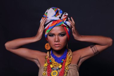 20 'li yaşlarda, Afrika Tepesi Kabilesi gibi giyinmiş Asyalı bir kadının vücudunun yarısını kapatın. Bronz tenli kız kendini güçlü hissediyor, gülümsüyor, renkli etnik aksesuarlarla mutlu, siyah arka plan