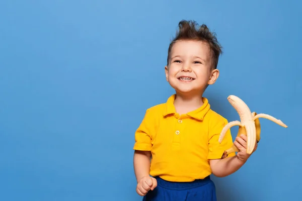 Feliz retrato de un niño en una camiseta amarilla sosteniendo un plátano pelado para la comida, plano de estudio aislado sobre un fondo azul —  Fotos de Stock