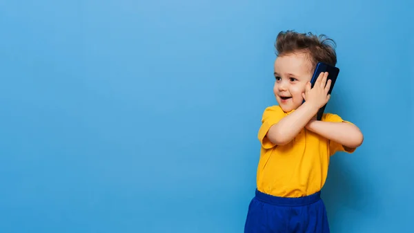 Un niño está hablando por un teléfono móvil. Foto del estudio sobre fondo azul. Espacio para texto —  Fotos de Stock