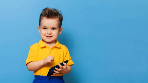 Un niño pequeño tiene un teléfono móvil en sus manos. Foto del estudio sobre fondo azul. Espacio para texto —  Fotos de Stock