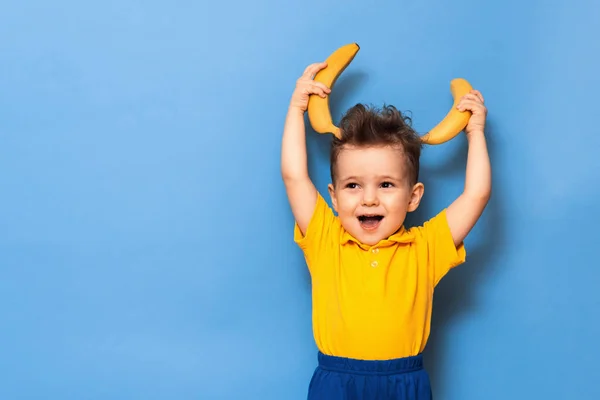 Feliz retrato de un niño con una sonrisa atractiva en una camiseta amarilla jugando sosteniendo un plátano, plano de estudio aislado sobre un fondo azul —  Fotos de Stock