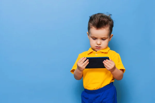 Un niño pequeño mira la pantalla de un teléfono móvil. Foto del estudio sobre fondo azul. —  Fotos de Stock