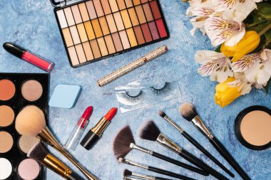 Dekoratif kozmetik seti, makyaj fırçaları ve mavi arka planda bir buket çiçek. Yukarıdan görünüm