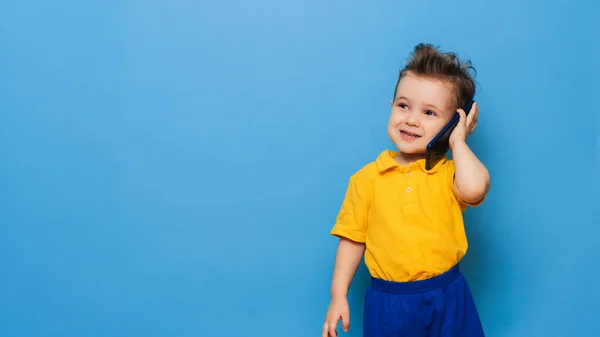Mały chłopiec rozmawia przez telefon komórkowy. Studio zdjęcie na niebieskim tle. Miejsce na tekst — Zdjęcie stockowe