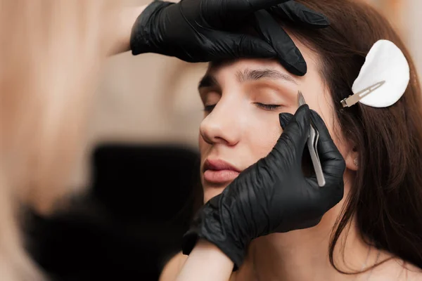 De cosmetoloog-brovist corrigeert en geeft vorm, plukt overtollige haren uit de wenkbrauwen met pincet in de schoonheidssalon. Professionele gezichtsverzorging — Stockfoto