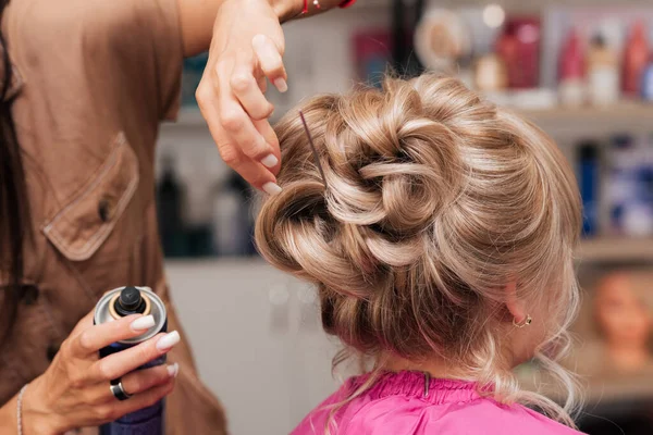 A menina-cabeleireiro faz o cliente um penteado de uma celebração. O cabeleireiro corrige tudo com spray de cabelo — Fotografia de Stock