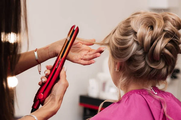 Uma menina-cabeleireiro faz um cabelo clientes para uma celebração com a ajuda de um alisador de ferro. — Fotografia de Stock