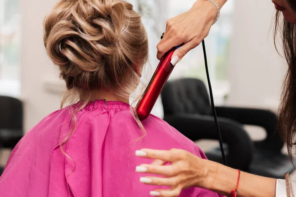 Uma menina-cabeleireiro faz um cabelo clientes para uma celebração com a ajuda de um alisador de ferro. — Fotografia de Stock