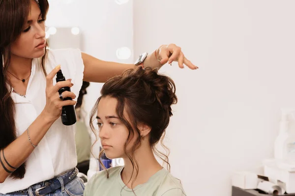 A filmar num salão de beleza. Um estilista mestre faz um penteado meninas de cabelos escuros com a ajuda de fixação com um spray de cabelo spray — Fotografia de Stock
