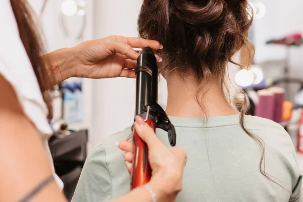 A filmar num salão de beleza. Um cabeleireiro faz um penteado para uma jovem garota de cabelos escuros com um ferro de ondulação — Fotografia de Stock