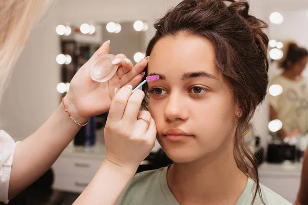 A filmar num salão de beleza. Um artista de maquiagem faz estilo de sobrancelha para uma jovem garota de cabelos escuros. — Fotografia de Stock