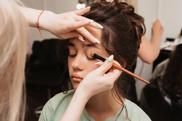 A filmar num salão de beleza. O maquiador aplica um pigmento brilhante aos olhos com uma escova macia. — Fotografia de Stock