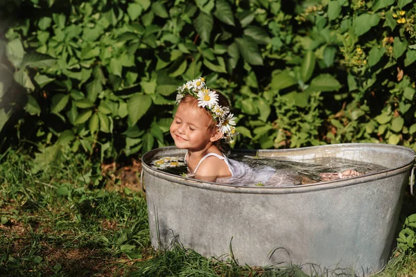 Un lindo bebé sonriente se está bañando en una bañera de pie en el jardín, en un día de verano soleado caliente —  Fotos de Stock