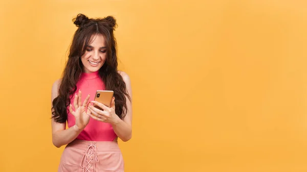 Uma menina segura seu smartphone em suas mãos e olha para a tela. A menina fica em um fundo amarelo. Uma cópia do espaço. Espaço para o seu texto. — Fotografia de Stock