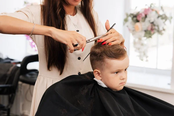 A filmar num salão de beleza. Um barbeiro corta um cabelo de garotinho com tesoura — Fotografia de Stock