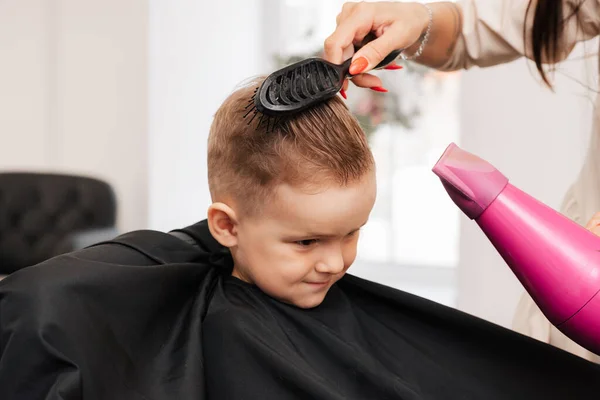 A filmar num salão de beleza. O cabeleireiro faz o estilo do menino com um secador de cabelo e um pente. — Fotografia de Stock