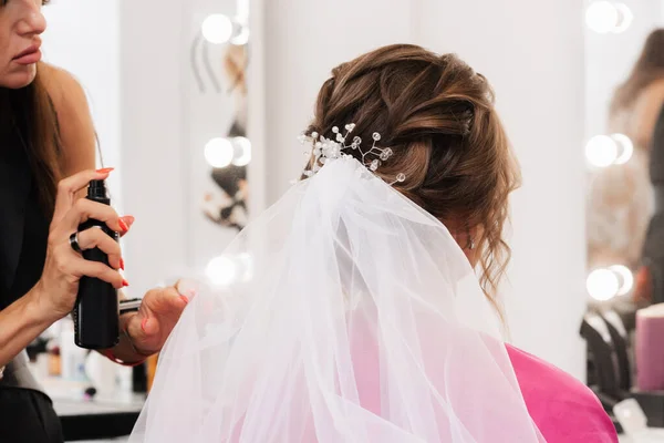 Um cabeleireiro faz um penteado elegante para estilizar uma noiva com um véu branco em seu cabelo no salão — Fotografia de Stock