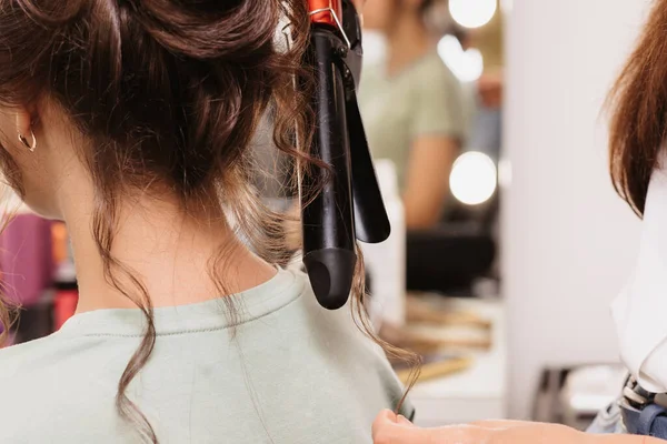 A filmar num salão de beleza. Um cabeleireiro faz um penteado para uma jovem garota de cabelos escuros com um ferro de ondulação — Fotografia de Stock