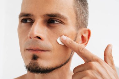 Beyaz bir arka planda, yüz kremi süren yakışıklı bir gencin yakın çekimi.