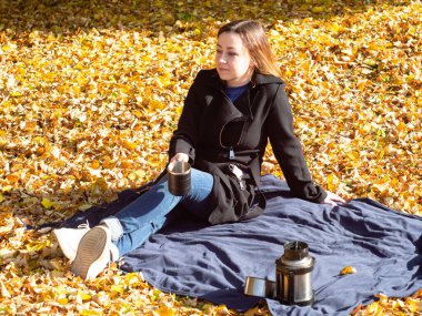 Elinde kupa olan genç bir kadın düşmüş yaprakların arasında bir battaniyenin üzerinde oturuyor. Park 'ta eğlence. Siyah yağmurluk ve kot pantolon giyen bir kadın..