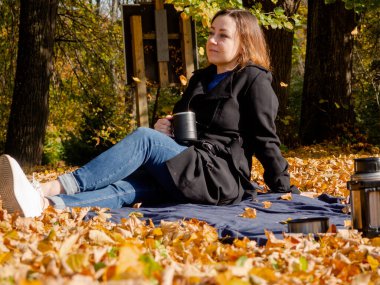 Elinde kupa olan genç bir kadın düşmüş yaprakların üzerinde battaniyenin üzerinde oturuyor. Siyah yağmurluk ve kot pantolon giyen bir kadın. Battaniyenin üzerinde piknik yaparken siyah termos kahve..