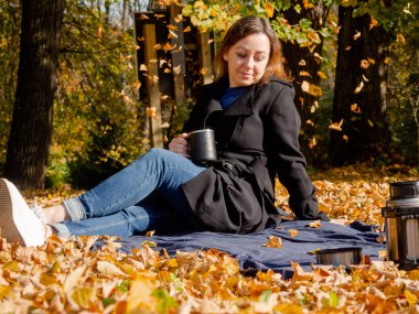 Elinde kupa olan genç bir kadın düşmüş yaprakların üzerinde battaniyenin üzerinde oturuyor. Siyah yağmurluk ve kot pantolon giyen bir kadın. Battaniyenin üzerinde piknik yaparken siyah termos kahve..