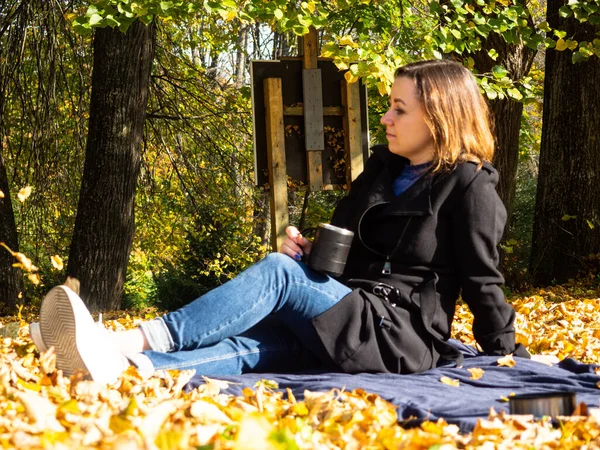 Elinde kupa olan genç bir kadın düşmüş yaprakların üzerinde battaniyenin üzerinde oturuyor. Siyah yağmurluk ve kot pantolon giyen bir kadın. Battaniyenin üzerinde piknik yaparken siyah termos kahve..