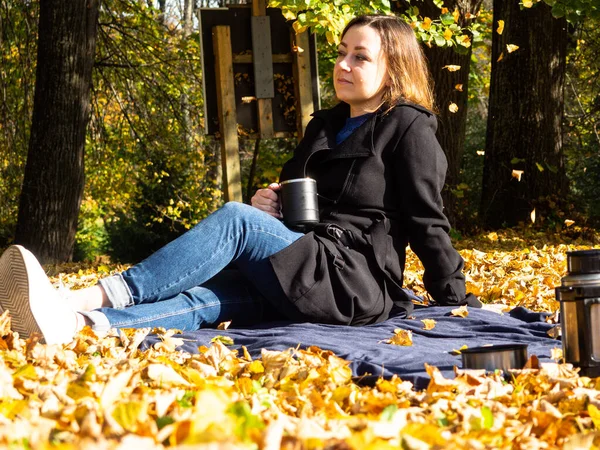 Elinde kupa olan genç bir kadın düşmüş yaprakların üzerinde battaniyenin üzerinde oturuyor. Siyah yağmurluk ve kot pantolon giyen bir kadın. Battaniyenin üzerinde piknik yaparken siyah termos kahve..