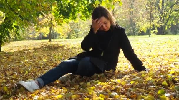 En ung kvinna rätar ut håret när hon sitter i en park på gula löv. promenader i höstparken — Stockvideo
