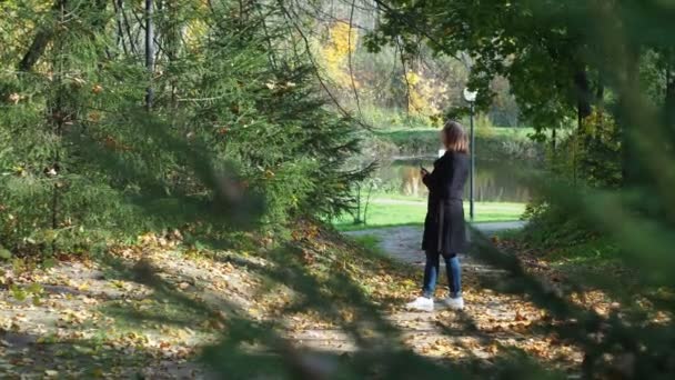 Bir sonbahar parkında elinde telefon olan siyah ceketli bir kız. Akıllı bir telefonla fotoğraf çekiyor. — Stok video