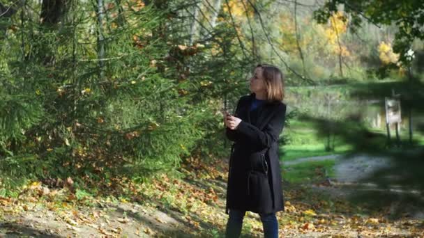 Bir sonbahar parkında elinde telefon olan siyah ceketli bir kız. Akıllı bir telefonla fotoğraf çekiyor. — Stok video