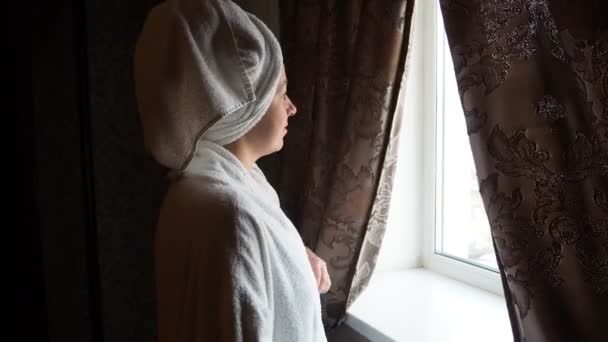 Jeune femme en peignoir regarde par la fenêtre dans une chambre d'hôtel. repos dans l'hôtel — Video