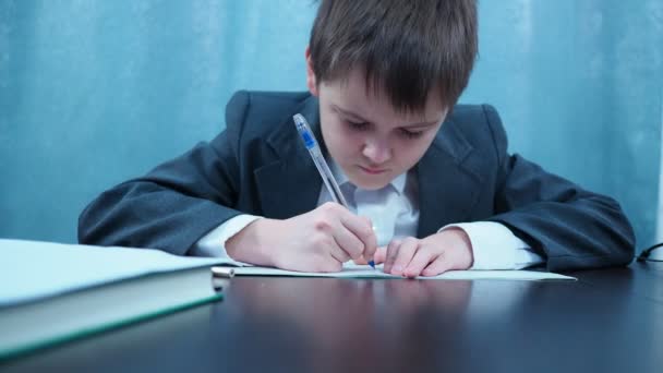 Мальчик в пиджаке и рубашке пишет в блокноте за столом — стоковое видео