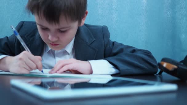 Ένα αγόρι με σακάκι γράφει σε ένα σημειωματάριο με ένα στυλό στο τραπέζι. Μπροστά του είναι ένα τάμπλετ με ακουστικά — Αρχείο Βίντεο